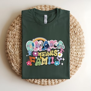 Puede incluir: Camiseta verde oscuro con el texto "Ohana Means Family" en una fuente retro colorida. El dise&ntilde;o incluye un arco&iacute;ris, flores y estrellas. La camiseta est&aacute; doblada sobre una estera circular tejida.