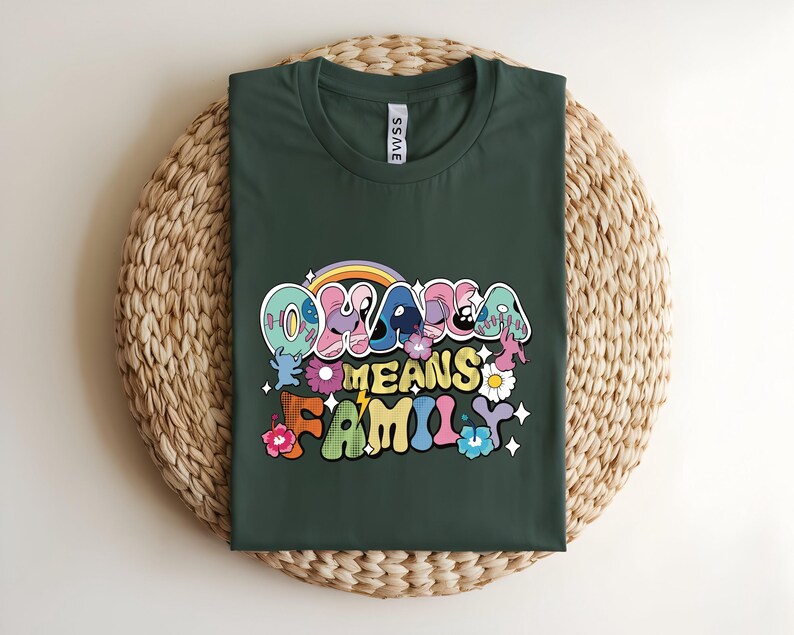 Puede incluir: Camiseta verde oscuro con el texto "Ohana Means Family" en una fuente retro colorida. El dise&ntilde;o incluye un arco&iacute;ris, flores y estrellas. La camiseta est&aacute; doblada sobre una estera circular tejida.