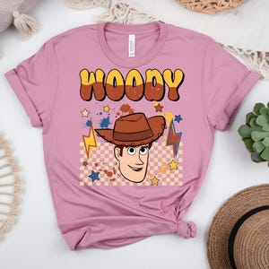 Puede incluir: Una camiseta rosa polvorienta con una imagen de dibujos animados de Woody de Toy Story. El dise&ntilde;o incluye el nombre "WOODY" en letras marrones y amarillas, una imagen de dibujos animados de Woody con un sombrero de vaquero marr&oacute;n y estrellas y rayos.