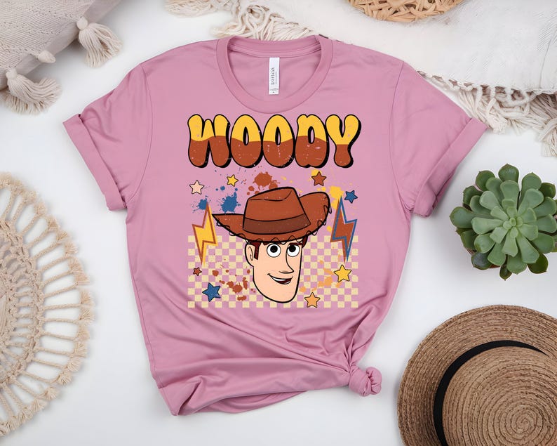 Puede incluir: Una camiseta rosa polvorienta con una imagen de dibujos animados de Woody de Toy Story. El dise&ntilde;o incluye el nombre "WOODY" en letras marrones y amarillas, una imagen de dibujos animados de Woody con un sombrero de vaquero marr&oacute;n y estrellas y rayos.
