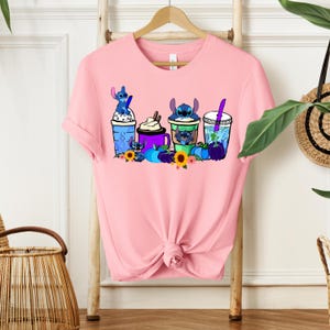 Puede incluir: Camiseta rosa claro con un dise&ntilde;o de personajes de Stitch en tazas de bebidas. El dise&ntilde;o incluye tazas azules, moradas y verdes con personajes de Stitch, girasoles y calabazas. La camiseta est&aacute; colgada en un perchero de madera.