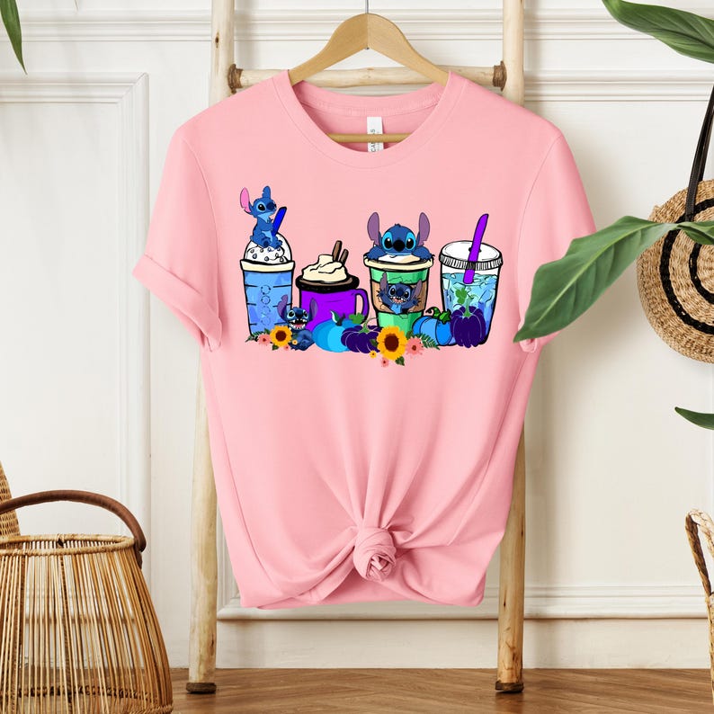 Puede incluir: Camiseta rosa claro con un dise&ntilde;o de personajes de Stitch en tazas de bebidas. El dise&ntilde;o incluye tazas azules, moradas y verdes con personajes de Stitch, girasoles y calabazas. La camiseta est&aacute; colgada en un perchero de madera.