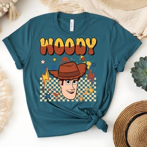 Puede incluir: Camiseta de color verde azulado con un gr&aacute;fico retro de Woody con un sombrero de vaquero marr&oacute;n. La palabra "WOODY" est&aacute; en una fuente de burbujas marr&oacute;n y amarilla. El dise&ntilde;o incluye estrellas, rayos y un fondo a cuadros.