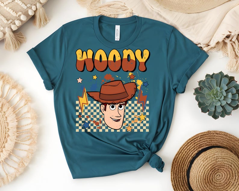 Puede incluir: Camiseta de color verde azulado con un gr&aacute;fico retro de Woody con un sombrero de vaquero marr&oacute;n. La palabra "WOODY" est&aacute; en una fuente de burbujas marr&oacute;n y amarilla. El dise&ntilde;o incluye estrellas, rayos y un fondo a cuadros.