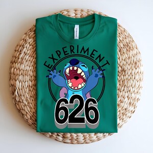 Pu&ograve; includere: Maglietta verde con un personaggio dei cartoni animati blu con la bocca aperta e le braccia alzate. Il testo "EXPERIMENT" &egrave; sopra e il numero "626" sotto.