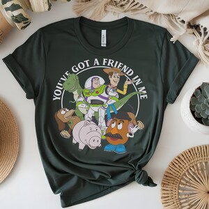 May include: Dark green t-shirt with a graphic of Toy Story characters. The design includes Woody, Buzz Lightyear, Rex, Mr. Potato Head, Hamm, and Slinky Dog, with the text "YOU'VE GOT A FRIEND IN ME" in a white circle.
