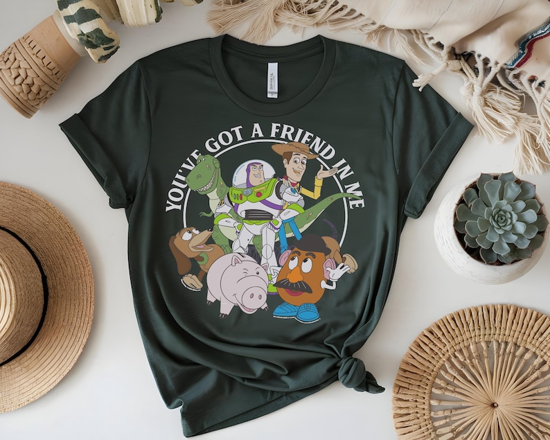 May include: Dark green t-shirt with a graphic of Toy Story characters. The design includes Woody, Buzz Lightyear, Rex, Mr. Potato Head, Hamm, and Slinky Dog, with the text "YOU'VE GOT A FRIEND IN ME" in a white circle.