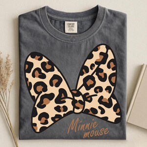 May include: A gray t-shirt featuring a large bow with a leopard print design. The bow is tan with brown spots. The words "Minnie mouse" are printed in a brown script font below the bow.
