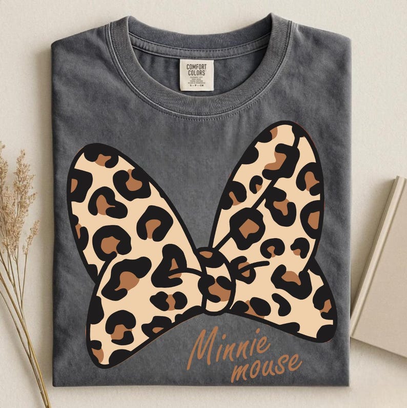 May include: A gray t-shirt featuring a large bow with a leopard print design. The bow is tan with brown spots. The words "Minnie mouse" are printed in a brown script font below the bow.