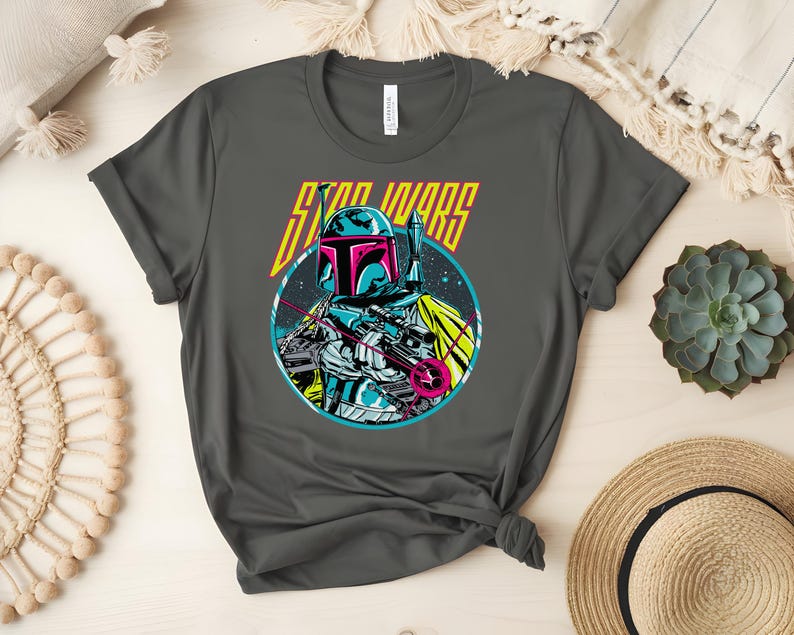 May include: Dark grey t-shirt with a vibrant graphic of Boba Fett from Star Wars. The design showcases a detailed illustration of the character with a blaster, set against a space backdrop, and the words "STAR WARS" in yellow.
