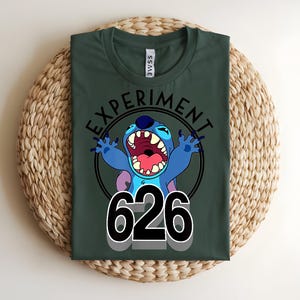 Pu&ograve; includere: T-shirt verde scuro con un personaggio dei cartoni animati blu, a bocca aperta, e la scritta "EXPERIMENT" sopra e "626" sotto. Il personaggio &egrave; all'interno di un cerchio nero.