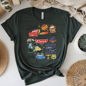 May include: Dark green t-shirt featuring a colorful graphic of characters and logos from the animated movie "Cars." The design includes Lightning McQueen, Mater, and other characters, along with the "Cars" logo and Route 66 signage.