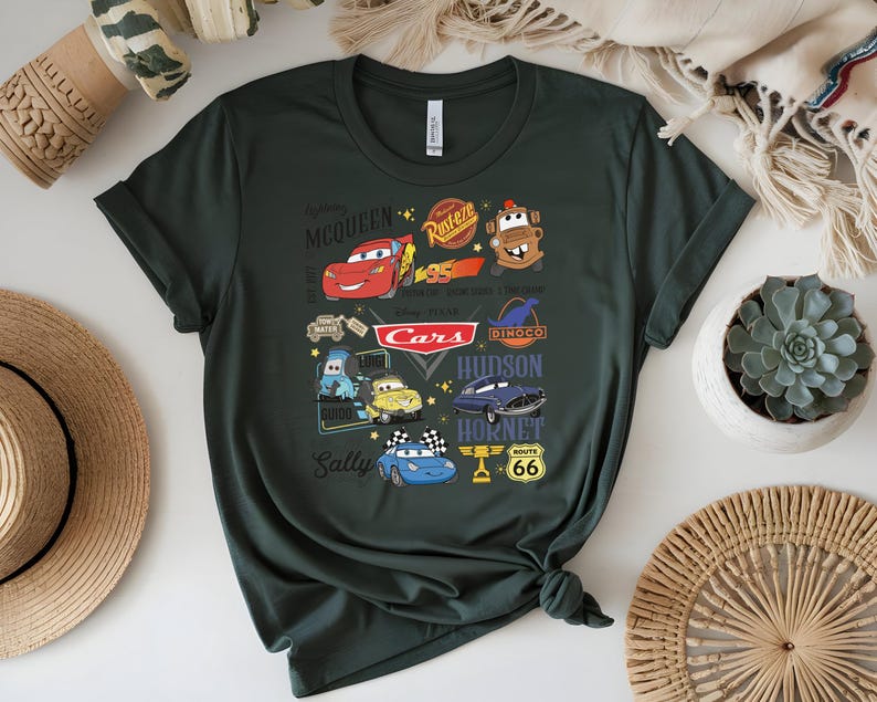 May include: Dark green t-shirt featuring a colorful graphic of characters and logos from the animated movie "Cars." The design includes Lightning McQueen, Mater, and other characters, along with the "Cars" logo and Route 66 signage.