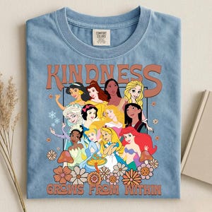 May include: Light blue Comfort Colors t-shirt featuring a graphic of various cartoon princesses with the word "KINDNESS" above them and the phrase "GROWS FROM WITHIN" below. The design includes flowers and mushrooms.