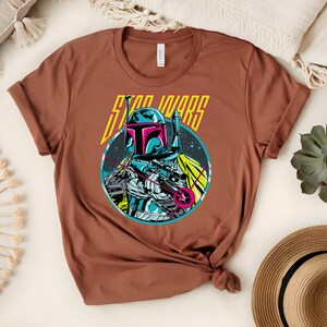 May include: A brown t-shirt with a graphic of a Star Wars character. The character is in a circular design with blue, pink, and white colours. The words "STAR WARS" are printed above the character in yellow.