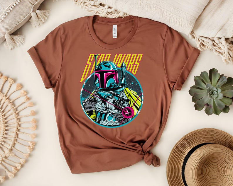May include: A brown t-shirt with a graphic of a Star Wars character. The character is in a circular design with blue, pink, and white colours. The words "STAR WARS" are printed above the character in yellow.