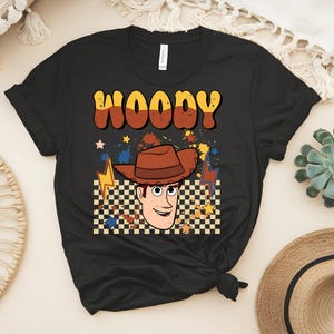 Puede incluir: Camiseta negra con un gr&aacute;fico de dibujos animados de Woody de Toy Story. El dise&ntilde;o incluye la cara de Woody, un sombrero de vaquero marr&oacute;n y el nombre "WOODY" en letras retro. El fondo tiene un patr&oacute;n de cuadros y rayos.