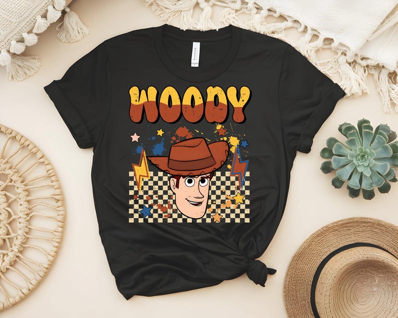 Puede incluir: Camiseta negra con un gr&aacute;fico de dibujos animados de Woody de Toy Story. El dise&ntilde;o incluye la cara de Woody, un sombrero de vaquero marr&oacute;n y el nombre "WOODY" en letras retro. El fondo tiene un patr&oacute;n de cuadros y rayos.