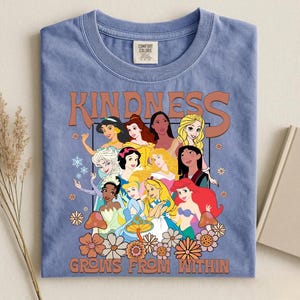 May include: A blue t-shirt featuring a graphic of various cartoon princesses with the word "KINDNESS" above them and the phrase "GROWS FROM WITHIN" below. The design includes flowers and mushrooms.