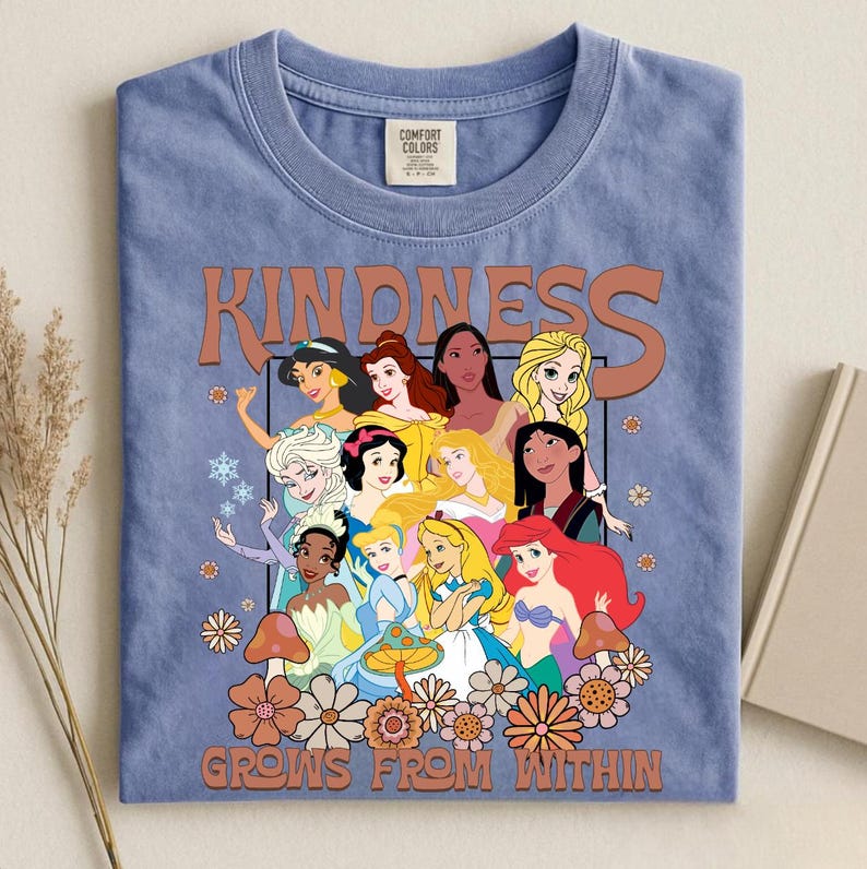 May include: A blue t-shirt featuring a graphic of various cartoon princesses with the word "KINDNESS" above them and the phrase "GROWS FROM WITHIN" below. The design includes flowers and mushrooms.