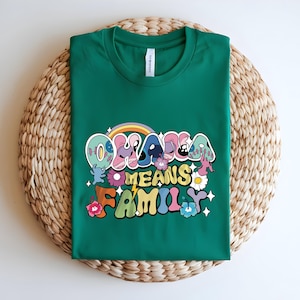 Puede incluir: Una camiseta verde con la frase "OHANA MEANS FAMILY" en letras coloridas de estilo retro. El dise&ntilde;o incluye un arco&iacute;ris, flores y estrellas. La camiseta se muestra sobre una estera redonda tejida.