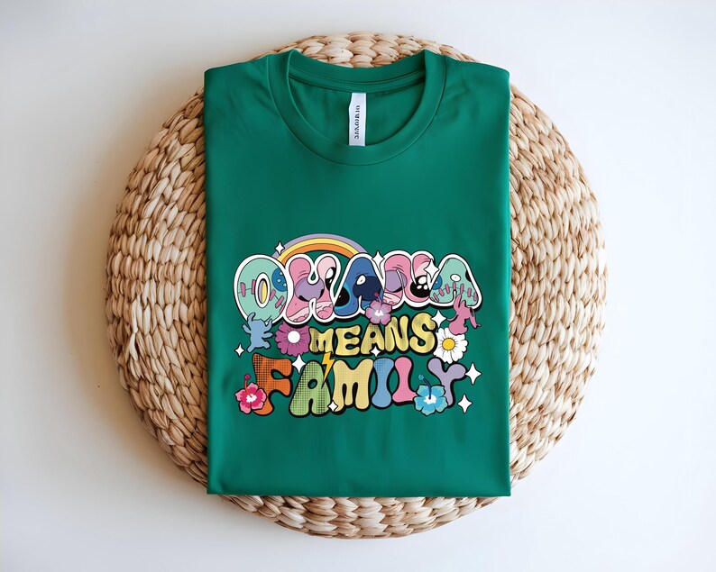 Puede incluir: Una camiseta verde con la frase "OHANA MEANS FAMILY" en letras coloridas de estilo retro. El dise&ntilde;o incluye un arco&iacute;ris, flores y estrellas. La camiseta se muestra sobre una estera redonda tejida.