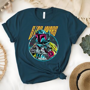 May include: Teal t-shirt with a Star Wars graphic. The design features Boba Fett in a circular image, with a helmet, blaster, and the words "STAR WARS" in yellow and pink. The shirt is made of a soft material.