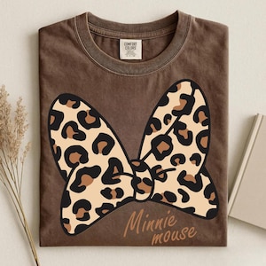 May include: Brown t-shirt featuring a large bow with a leopard print design. The bow is tan with black and brown spots. The words "Minnie mouse" are written in a brown script font below the bow. The shirt is folded.