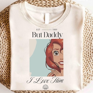 May include: Cream-colored t-shirt featuring the text "But Daddy" above a cartoon image of a red-haired character, with the words "I Love Him" below. A seashell graphic is at the bottom. The shirt has a round neckline.