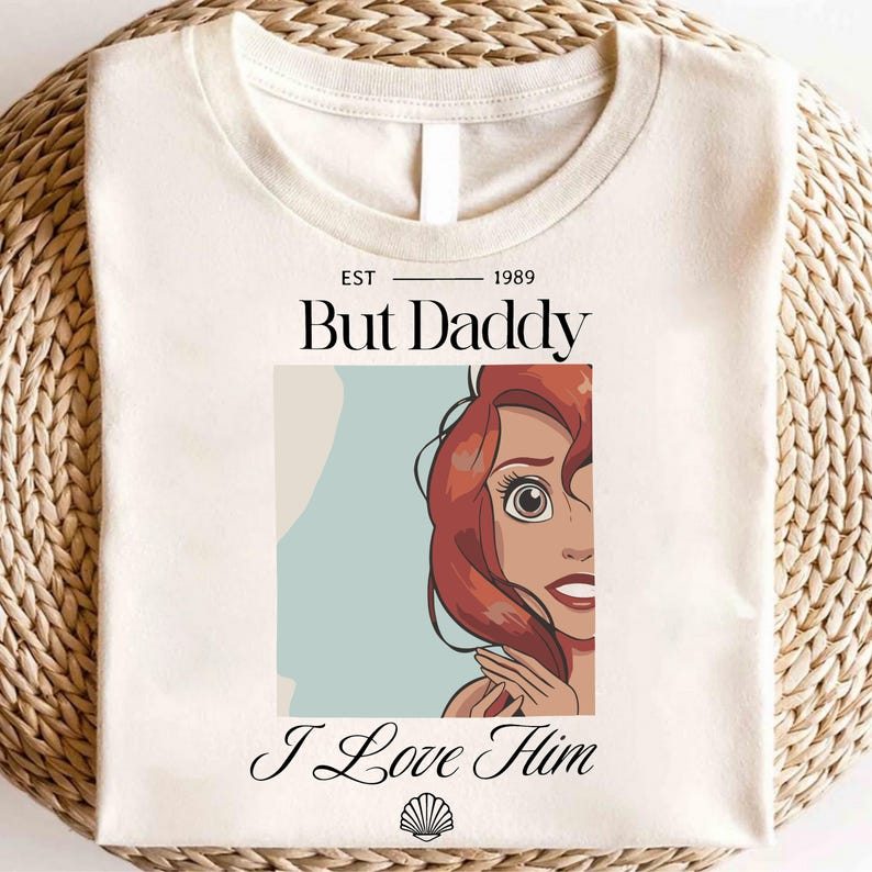 May include: Cream-colored t-shirt featuring the text "But Daddy" above a cartoon image of a red-haired character, with the words "I Love Him" below. A seashell graphic is at the bottom. The shirt has a round neckline.