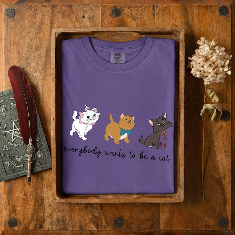 May include: A lilac t-shirt with three cartoon cats, each with a bow or bow tie. The phrase "everybody wants to be a cat" is printed below. The shirt is presented in a wooden tray.