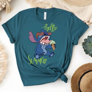 K&ouml;nnte beinhalten: T&uuml;rkisfarbenes T-Shirt mit einer Comicfigur, die eine Sonnenbrille tr&auml;gt und eine Eist&uuml;te h&auml;lt. Der Text "hello summer" ist in gr&uuml;ner Schreibschrift geschrieben. Die Figur ist blau mit gro&szlig;en Ohren und offenem Mund.
