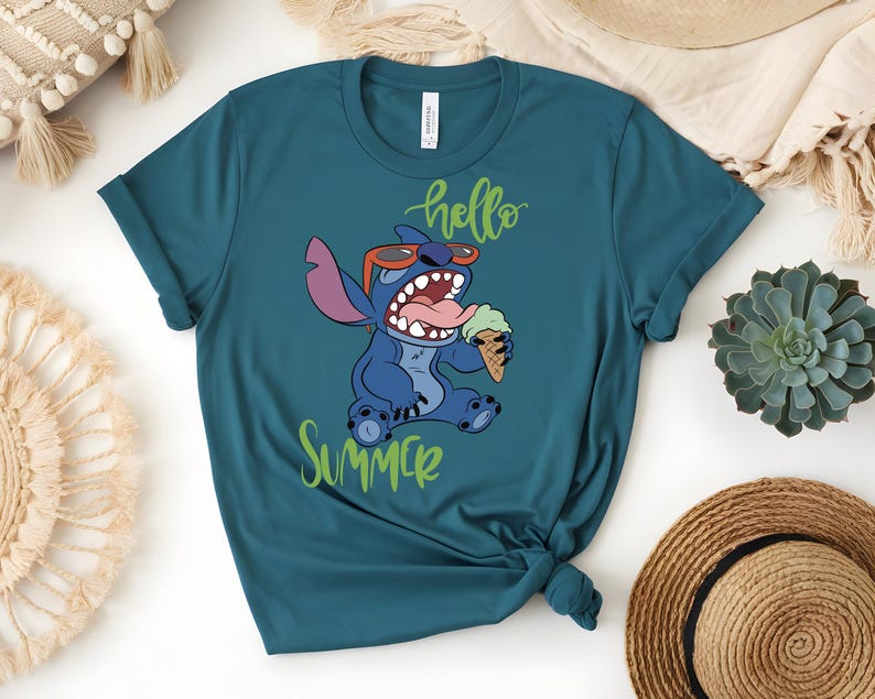 K&ouml;nnte beinhalten: T&uuml;rkisfarbenes T-Shirt mit einer Comicfigur, die eine Sonnenbrille tr&auml;gt und eine Eist&uuml;te h&auml;lt. Der Text "hello summer" ist in gr&uuml;ner Schreibschrift geschrieben. Die Figur ist blau mit gro&szlig;en Ohren und offenem Mund.