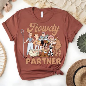 May include: A rust-colored t-shirt featuring a graphic of cartoon characters with the text "Howdy Partner" in a retro font. The design includes characters like Woody, Buzz Lightyear, and Mickey Mouse, evoking a Western theme.