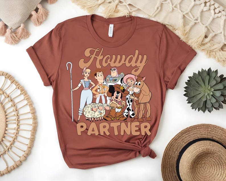 May include: A rust-colored t-shirt featuring a graphic of cartoon characters with the text "Howdy Partner" in a retro font. The design includes characters like Woody, Buzz Lightyear, and Mickey Mouse, evoking a Western theme.