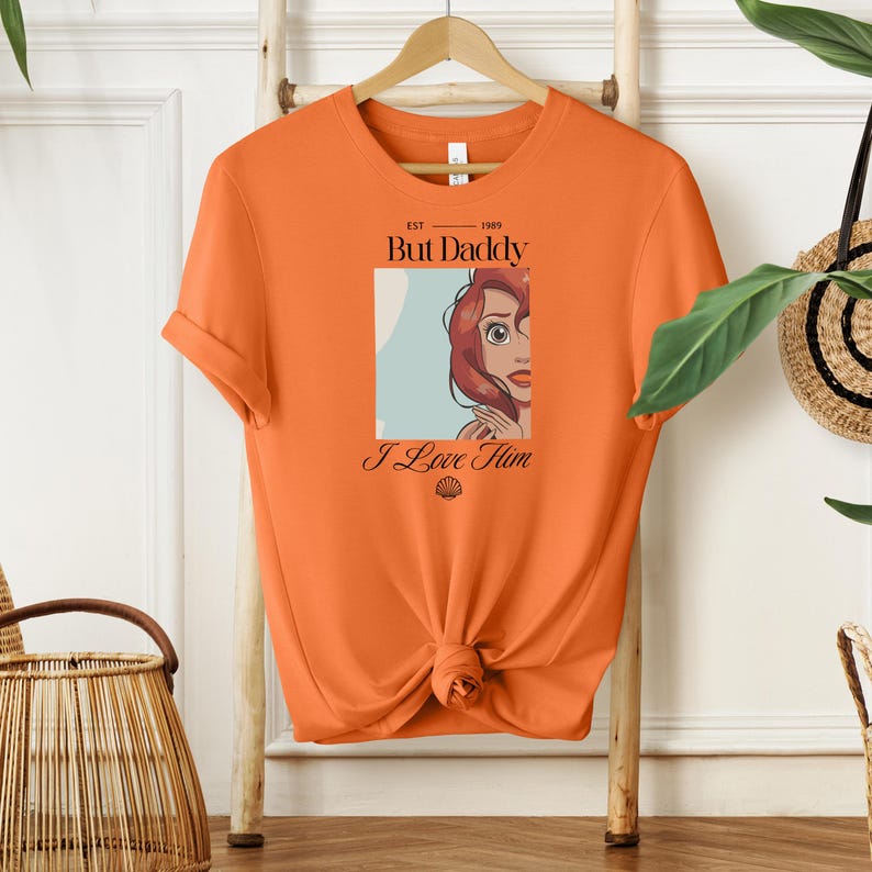 May include: Orange t-shirt with a graphic of a cartoon woman with red hair and the text "But Daddy I Love Him" in black. The shirt is knotted at the waist and hangs on a wooden hanger.