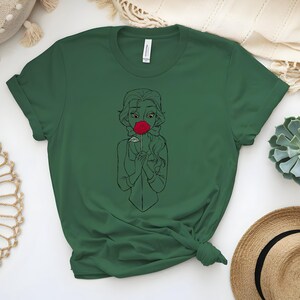 May include: Dark green t-shirt featuring a line drawing of a woman holding a red rose to her face. The woman has long hair and is wearing a simple dress. The t-shirt is made of a soft material and has short sleeves.