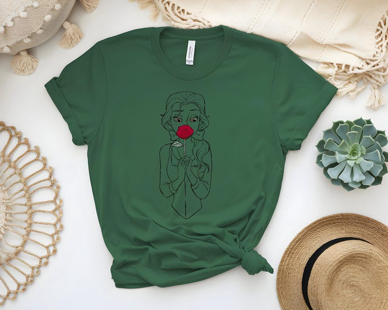 May include: Dark green t-shirt featuring a line drawing of a woman holding a red rose to her face. The woman has long hair and is wearing a simple dress. The t-shirt is made of a soft material and has short sleeves.