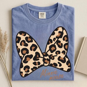 May include: A blue t-shirt with a large bow design. The bow features a tan leopard print with brown spots. The words "Minnie mouse" are printed in a brown script font below the bow. The shirt has a Comfort Colors tag.
