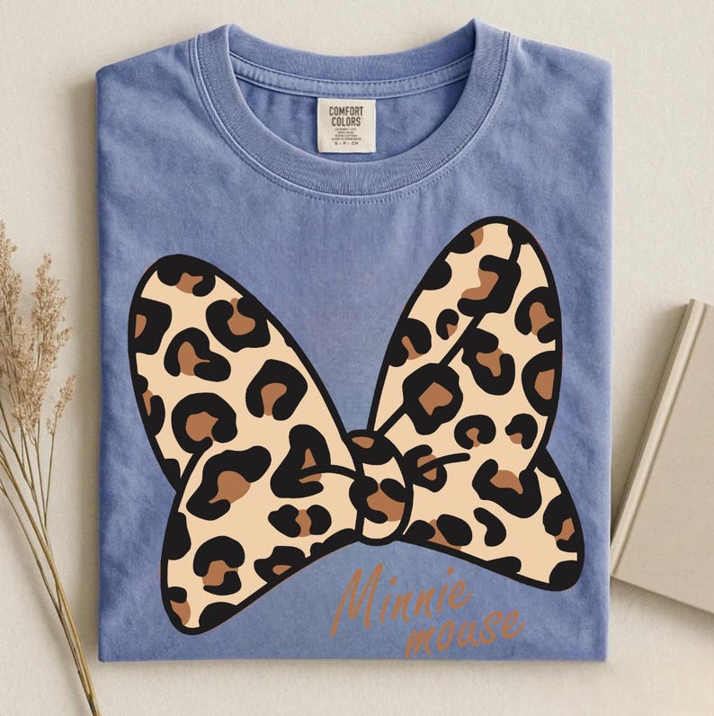 May include: A blue t-shirt with a large bow design. The bow features a tan leopard print with brown spots. The words "Minnie mouse" are printed in a brown script font below the bow. The shirt has a Comfort Colors tag.
