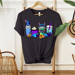 Puede incluir: Camiseta negra con un gr&aacute;fico colorido de personajes de Stitch en varias bebidas. El dise&ntilde;o incluye bebidas azules, moradas y verdes, girasoles y calabazas. La camiseta est&aacute; colgada en un perchero de madera.