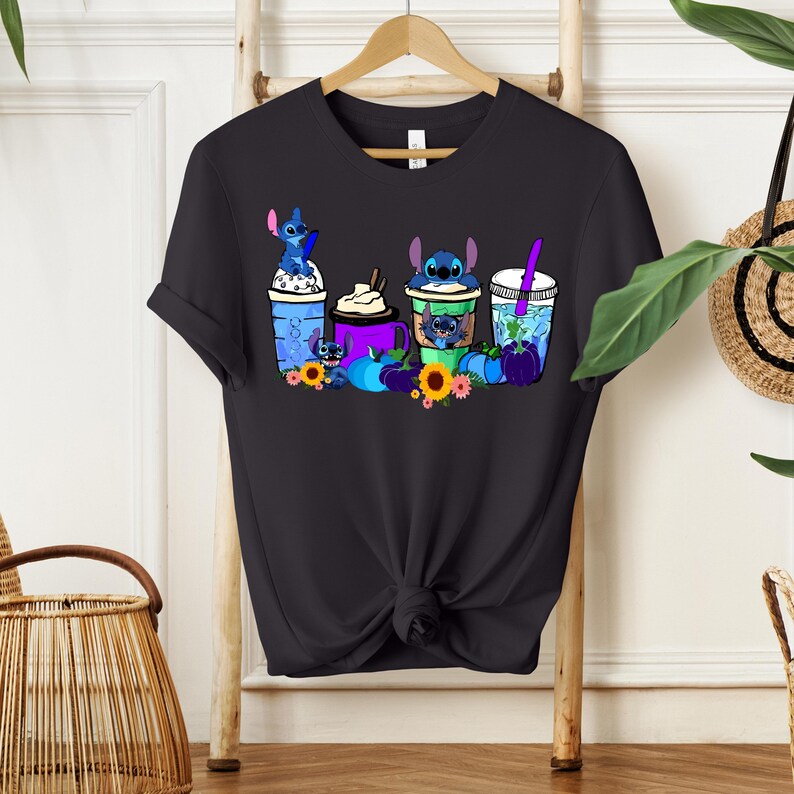 Puede incluir: Camiseta negra con un gr&aacute;fico colorido de personajes de Stitch en varias bebidas. El dise&ntilde;o incluye bebidas azules, moradas y verdes, girasoles y calabazas. La camiseta est&aacute; colgada en un perchero de madera.
