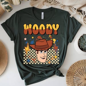 Puede incluir: Camiseta verde oscuro con una ilustraci&oacute;n de dibujos animados de Woody de Toy Story. El dise&ntilde;o incluye el nombre "WOODY" en naranja y amarillo, un sombrero de vaquero marr&oacute;n y un patr&oacute;n de cuadros. Estrellas y rayos completan el dise&ntilde;o.