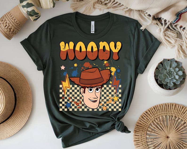 Puede incluir: Camiseta verde oscuro con una ilustraci&oacute;n de dibujos animados de Woody de Toy Story. El dise&ntilde;o incluye el nombre "WOODY" en naranja y amarillo, un sombrero de vaquero marr&oacute;n y un patr&oacute;n de cuadros. Estrellas y rayos completan el dise&ntilde;o.