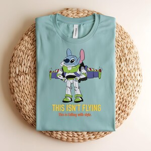 May include: A light blue t-shirt featuring a cartoon character, Stitch, dressed as Buzz Lightyear. The graphic includes the text "THIS ISN'T FLYING" and "this is falling with style." The shirt is displayed on a woven, circular mat.