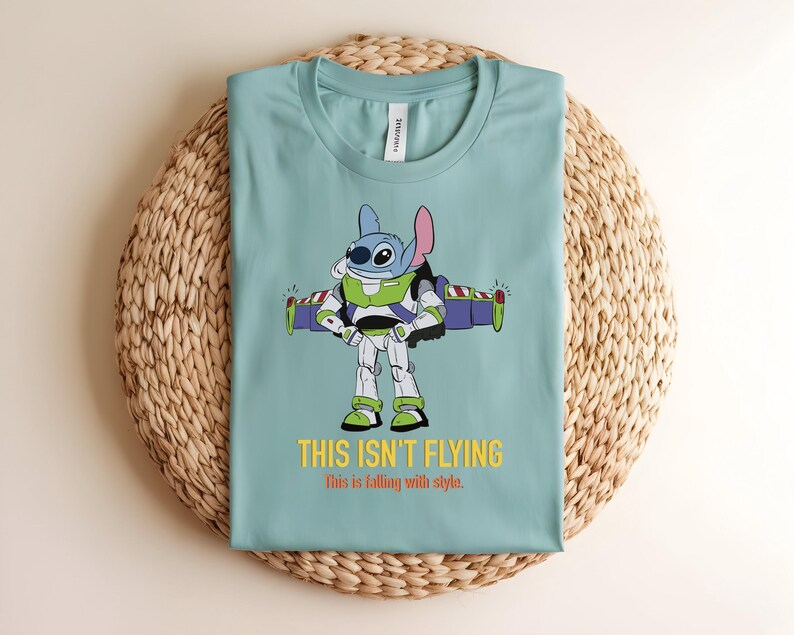 May include: A light blue t-shirt featuring a cartoon character, Stitch, dressed as Buzz Lightyear. The graphic includes the text "THIS ISN'T FLYING" and "this is falling with style." The shirt is displayed on a woven, circular mat.
