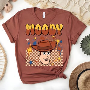 Puede incluir: Una camiseta de color &oacute;xido con un gr&aacute;fico de Woody de Toy Story. El dise&ntilde;o incluye el nombre "WOODY" en una fuente retro, una imagen de dibujos animados de Woody con un sombrero de vaquero, estrellas y rayos. El fondo tiene un patr&oacute;n de cuadros.