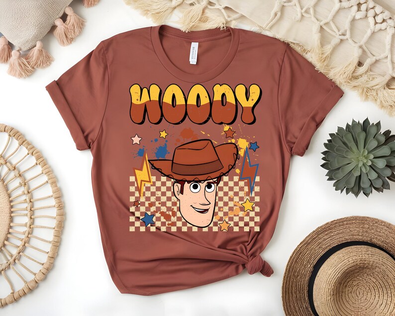 Puede incluir: Una camiseta de color &oacute;xido con un gr&aacute;fico de Woody de Toy Story. El dise&ntilde;o incluye el nombre "WOODY" en una fuente retro, una imagen de dibujos animados de Woody con un sombrero de vaquero, estrellas y rayos. El fondo tiene un patr&oacute;n de cuadros.