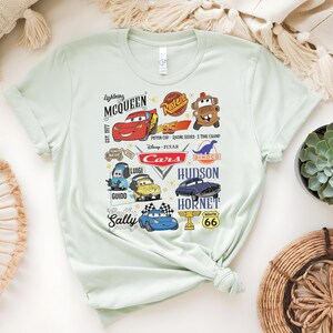 May include: Light green t-shirt featuring various characters and logos from the Disney Pixar Cars movie. The design includes Lightning McQueen, Mater, Sally, and other characters, along with the text "Cars" and "Route 66".