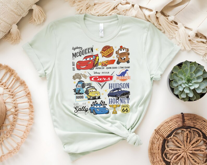 May include: Light green t-shirt featuring various characters and logos from the Disney Pixar Cars movie. The design includes Lightning McQueen, Mater, Sally, and other characters, along with the text "Cars" and "Route 66".